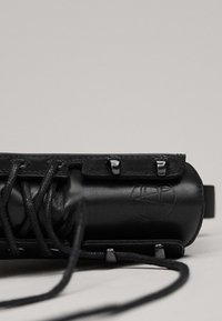 Gros plan de la partie supérieure de la botte en cuir noir avec des lacets passés dans des œillets métalliques sur un fond neutre.