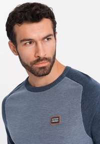 Sudadera de punto con textura acanalada, tonos en degradado de azul, cuello redondo y un pequeño parche con el logo en el pecho.