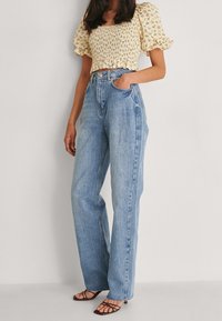 Top court floral avec manches bouffantes en jaune ; jeans bleu clair taille haute et ample ; mannequin portant des sandales brunes à brides ; textures lisses.