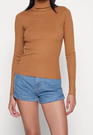 Donna con un maglione aderente a coste marrone con collo alto e pantaloncini di jeans blu risvoltati su uno sfondo semplice.