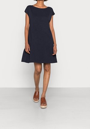 Robe bleu marine à manches courtes avec une coupe décontractée et un ourlet évasé. Fabriquée en tissu léger, accompagnée de sandales à glisser tissées de couleur marron.