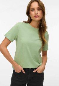 Γυναίκα με μακριά καστανά μαλλιά, που φοράει ανοιχτό πράσινο μπλουζάκι με κοντά μανίκια και μαύρο παντελόνι, στέκεται με τα χέρια στις τσέπες μπροστά σε λευκό φόντο.