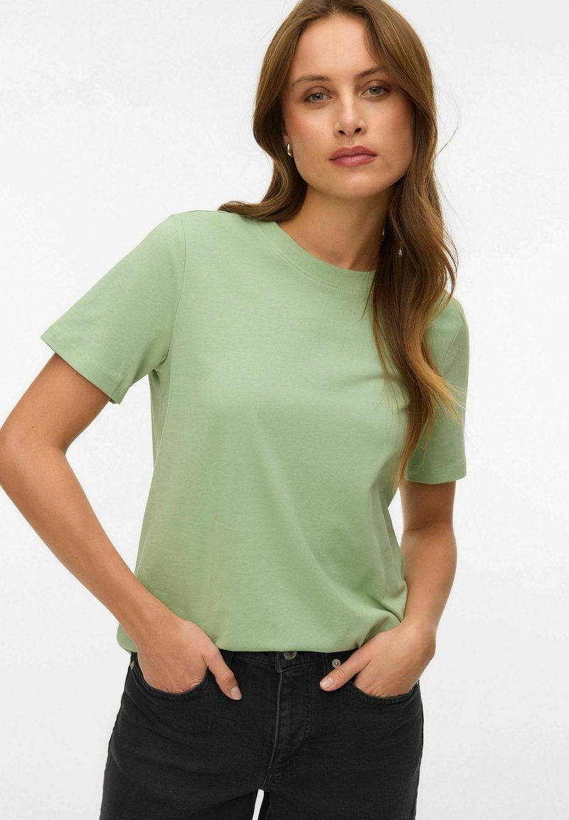 Γυναίκα με μακριά καστανά μαλλιά, που φοράει ανοιχτό πράσινο μπλουζάκι με κοντά μανίκια και μαύρο παντελόνι, στέκεται με τα χέρια στις τσέπες μπροστά σε λευκό φόντο.