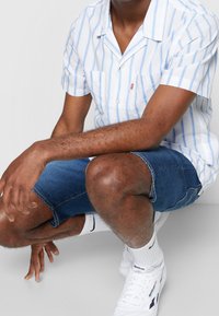 Camisa de manga corta, con cuello, en blanco con rayas azules, combinada con pantalones cortos de denim azul. Zapatillas blancas y calcetines de longitud media completan el look.