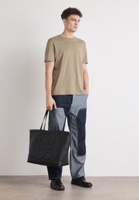 Lysebrun bomulds t-shirt, mørkeblå og grå patchwork jeans, og en sort tekstil tote med præget "HUGO." Sorte slip-on sko.