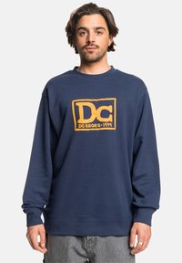 Navy blue sweatshirt made of cotton blend, featuring a large orange DC logo and text. Crew neck and long sleeves with ribbed cuffs.