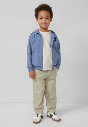 Kind staat en draagt een blauwe hoodie met ritssluiting, een wit T-shirt, beige broek en witte sneakers met zwarte en bruine accenten.