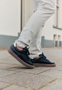 Sneaker in pelle nera e suede con suola in gomma marrone, caratterizzati da un design con lacci e accenti bianchi sul tallone in contrasto.