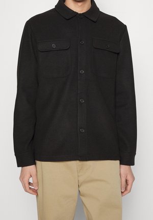 Mann trägt ein schwarzes Hemdjacke mit zwei geknöpften Brusttaschen und beige Hose vor einfarbigem Hintergrund.