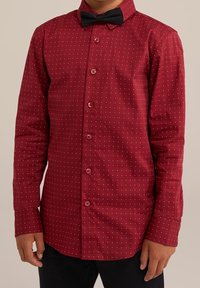 Röd skjorta med knäppning fram, vita prickar, långa ärmar och en svart fluga. Har en krage och matchande röda knappar. Slät textur.