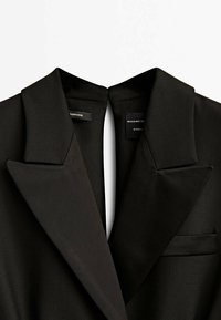 Svart blazer med slagkrage, med en ren design, jämn textur och en enda bröstficka. Inre etiketter synliga.