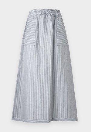 Žena stojaca pri jednoduchej stene, oblečená v svetlomodrú košeľu na gombíky, dlhú bielo-modro prúžkovanú sukňu a čierne sandále.