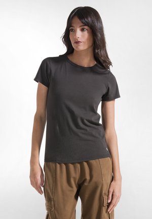 Młoda kobieta o długich ciemnych włosach, ubrana w czarny t-shirt z krótkim rękawem i brązowe spodnie typu cargo, stoi na jasnym, jednolitym tle.