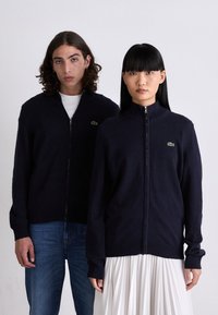 Maglione con zip navy, colletto e polsini a coste, con piccolo logo verde. Il modello abbina il maglione a jeans blu; il secondo modello indossa una gonna plissettata.