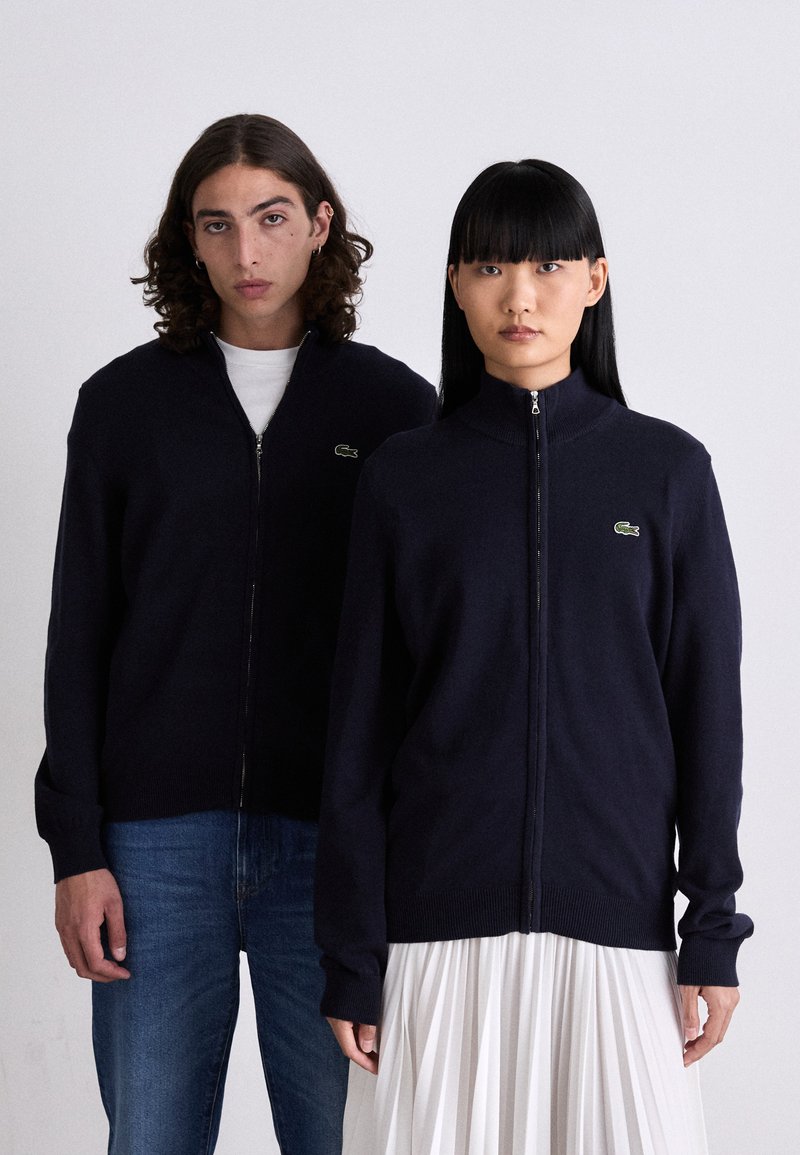 Maglione con zip navy, colletto e polsini a coste, con piccolo logo verde. Il modello abbina il maglione a jeans blu; il secondo modello indossa una gonna plissettata.