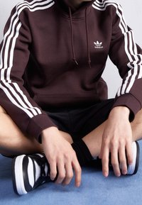 Sudadera marrón con diseño de tres rayas blancas en las mangas, capucha con cordón y pequeño logo bordado. Se lleva con pantalones cortos negros y zapatillas.
