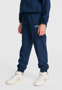 Marina byxor i texturerad elastisk tyg med resår vid anklarna, med "hummel"-logotyp på låret. Parat med vita högskafflade sneakers.