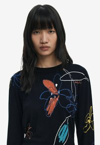 Pull en tricot noir avec une broderie florale colorée en orange, bleu, jaune et vert. Manches longues et décolleté rond.