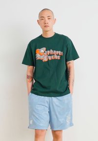 Grünes T-Shirt mit einem grafischen Druck, der eine Cartoonfigur und den Text "Carhartt Bucks" zeigt. Hellblaue Shorts mit dezenten Mustern.