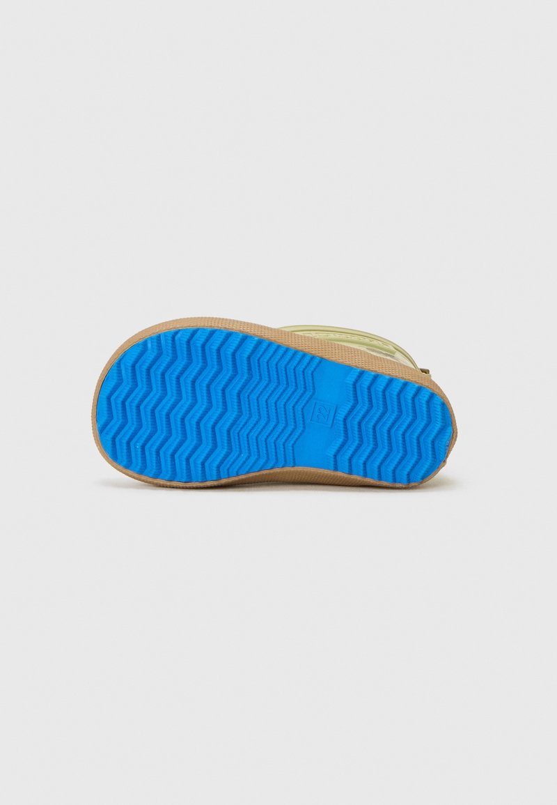 Une chaussure beige et bleue unique avec une semelle à motif zigzag texturé, orientée vers le haut, sur un fond gris clair uni.