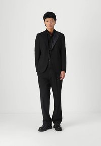 Black suit with a satin lapel, paired with a black shirt and trousers. Smooth fabric with a tailored fit, finished with black footwear.