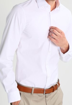En man som bär en vit långärmad skjorta med knappar, instoppad i beige byxor med ett brunt skinnbälte, justerar skjortan vid kragen.