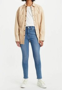 Camisa chaqueta de gran tamaño en color beige con dos bolsillos en el pecho, llevada sobre una camiseta blanca y unos vaqueros ajustados azules, completada con zapatillas blancas.