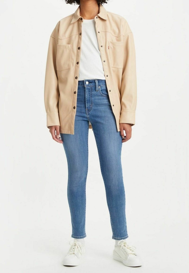 Camisa chaqueta de gran tamaño en color beige con dos bolsillos en el pecho, llevada sobre una camiseta blanca y unos vaqueros ajustados azules, completada con zapatillas blancas.