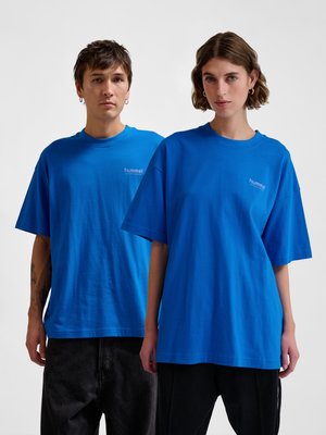 Twee volwassenen dragen identieke felblauwe oversized hummel sport-T-shirts, staand zij aan zij tegen een effen achtergrond.