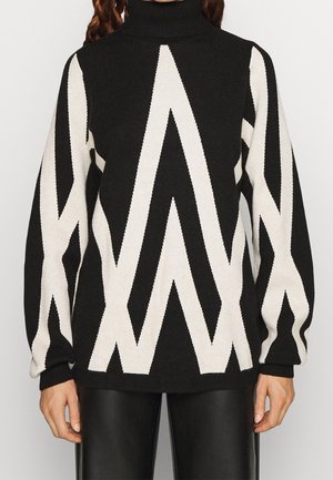 Femme portant un pull à col roulé noir avec de larges motifs zigzag blancs et de longues manches bouffantes, associé à un pantalon en cuir noir.