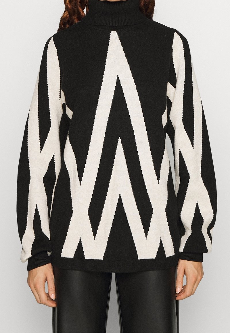 Femme portant un pull à col roulé noir avec de larges motifs zigzag blancs et de longues manches bouffantes, associé à un pantalon en cuir noir.