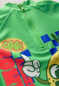 Rash guard verde a maniche corte con zip frontale, stampe colorate con testi e grafiche giocose, tessuto morbido e colori vivaci.