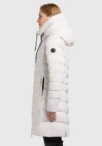 Woman wearing a long, white padded winter coat with a hood, side zipper pockets, and a black logo patch on the sleeve, standing sideways.
