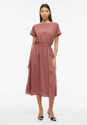 Vrouw die een mauve midi-jurk draagt met korte mouwen, een elastische tailleband en zwarte open teen hoge hakken, staand op een witte achtergrond.