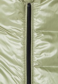 Puffjacke in hellgrün, mit einem zentralen schwarzen Reißverschluss, glatter Textur und glänzendem Finish mit dezenten Falten.