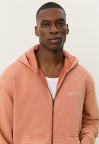 Hooded zip-up dukserica u izblijedjeloj koralj boji, napravljena od mekanog materijala, s prednjim džepom i izvezenim detaljem "LES DEUX".