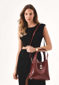 Borsa a tracolla in pelle bordeaux con finitura lucida, design strutturato e dettagli in oro. Presenta una singola tracolla e una chiusura superiore con fermaglio.