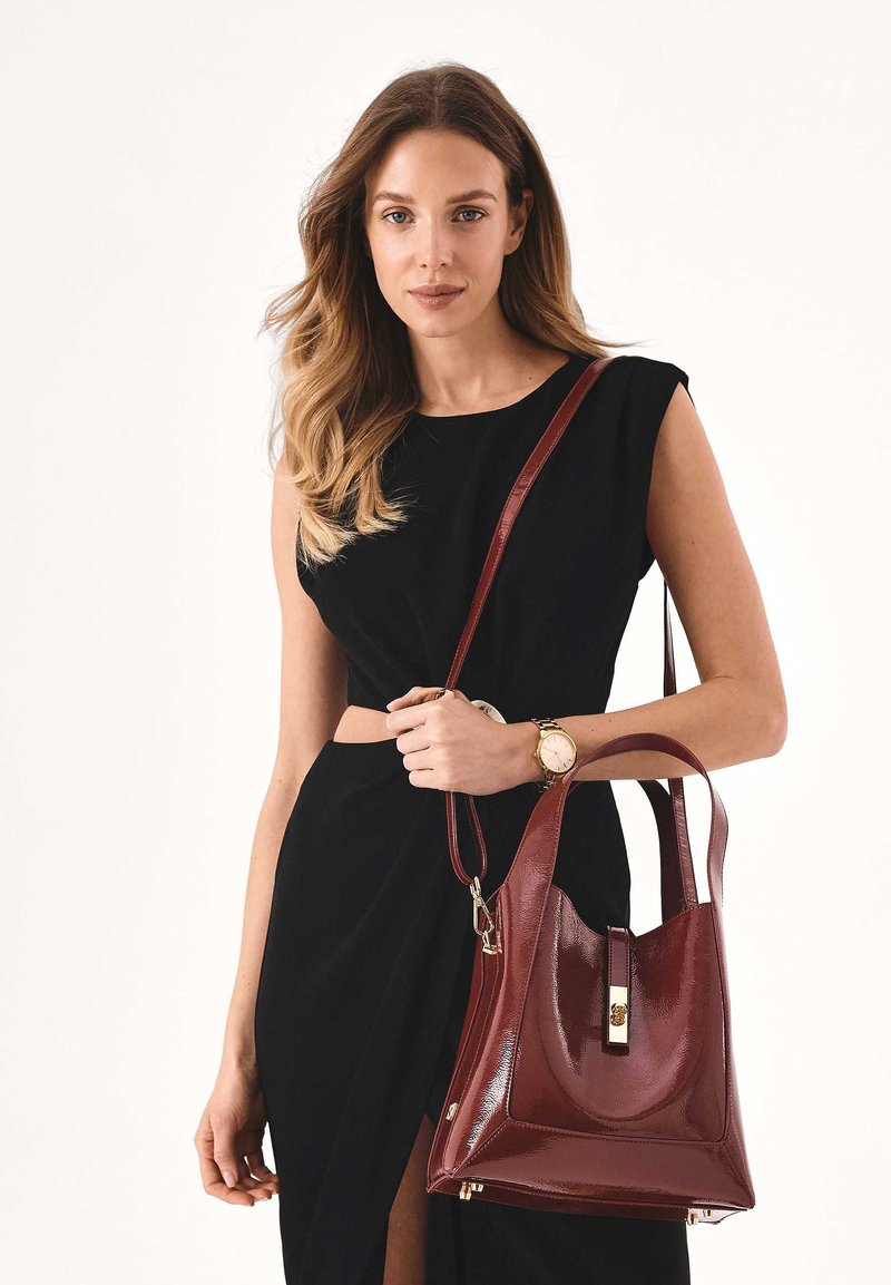 Borsa a tracolla in pelle bordeaux con finitura lucida, design strutturato e dettagli in oro. Presenta una singola tracolla e una chiusura superiore con fermaglio.