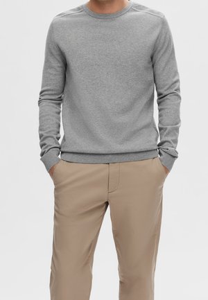 Mann in hellgrauem Langarmshirt mit Rundhalsausschnitt und beigen Hosen, die die Hände in den Taschen hat, vor einfachem Hintergrund.