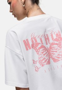 T-shirt bianca oversize con un grande grafico di una farfalla rosa e il testo "Good For Nothing" sul retro. Maniche corte e vestibilità rilassata.