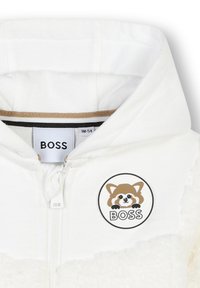 Bijela zip-sveta jakna s teksturiranim flis tijelom, glatkim kontrastnim kapuljačom i kružnom oznakom s crtanom rakunom i "BOSS" logom.