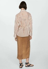 Camicia beige a righe con linee rosse e vita con laccetti, abbinata a una gonna di raso marrone e sandali neri. Sfondo bianco semplice.