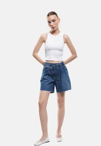 Nuori nainen seisoo käsivarsien takana, päällään valkoinen hihaton crop-top, epäsymmetriset siniset farkkushortsit ja valkoiset ballerinat.