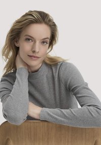 Grau gerippter Langarmshirt mit hohem Kragen. Der Stoff wirkt weich und strukturiert. Hände ruhen auf einem Holzstuhl mit glatter Oberfläche.