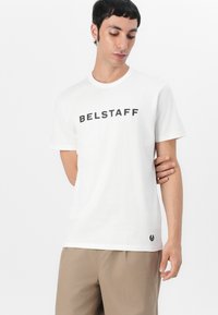 Fehér pamut póló, amelyen merész fekete "BELSTAFF" felirat található. Egyszerű, kerek nyakú dizájn, rövid ujjakkal és kis logó részlettel az alsó szegélyen.
