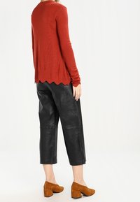 Roter Cardigan mit langen Ärmeln und gewelltem Saum, kombiniert mit schwarzen, kurzen Lederhosen und braunen, klobigen Absatzschuhen.