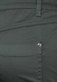 Mörkgröna byxor har en slät tygtyp med synliga stygn och en metallknapp som accent på framfickan. Texturen verkar mjuk.