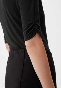 Zwarte, getailleerde top met driekwartmouwen, met een geplooid detail bij de manchetten, gecombineerd met hoge zwarte broek.