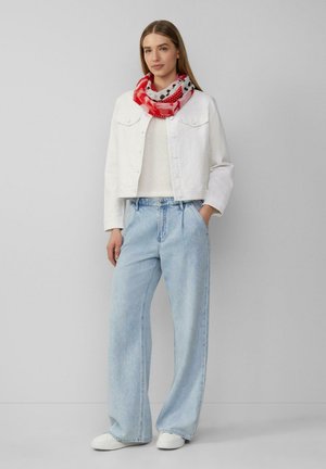 Vrouw draagt een witte cropped jas, crèmekleurige trui, lichtblauwe wijde jeans, witte sneakers en een rood patroon sjaal, staand tegen een grijze achtergrond.