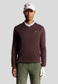 Maglione a maglia marrone con scollo a V, camicia bianca con colletto sotto, abbinato a pantaloni verde oliva e un cappellino bianco con un logo giallo.